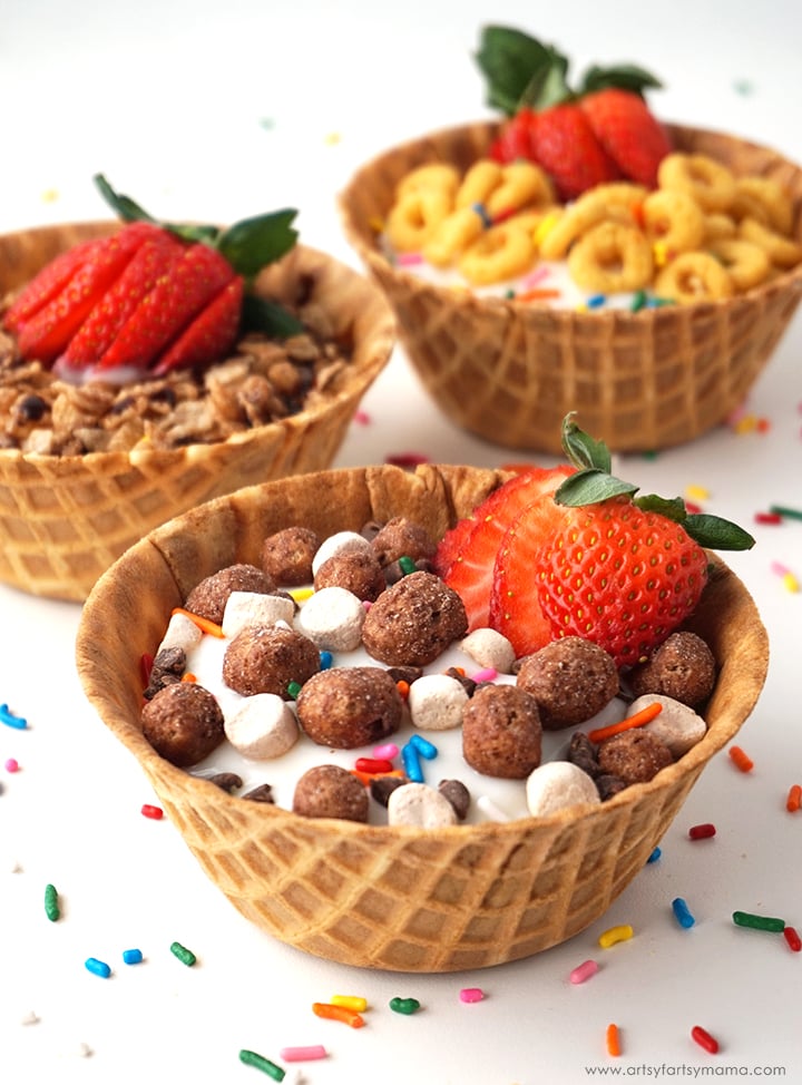 Three waffle bowls filled with cereal, yoghurt, and strawberries, topped with colourful sprinkles, are arranged on a white surface with scattered sprinkles.