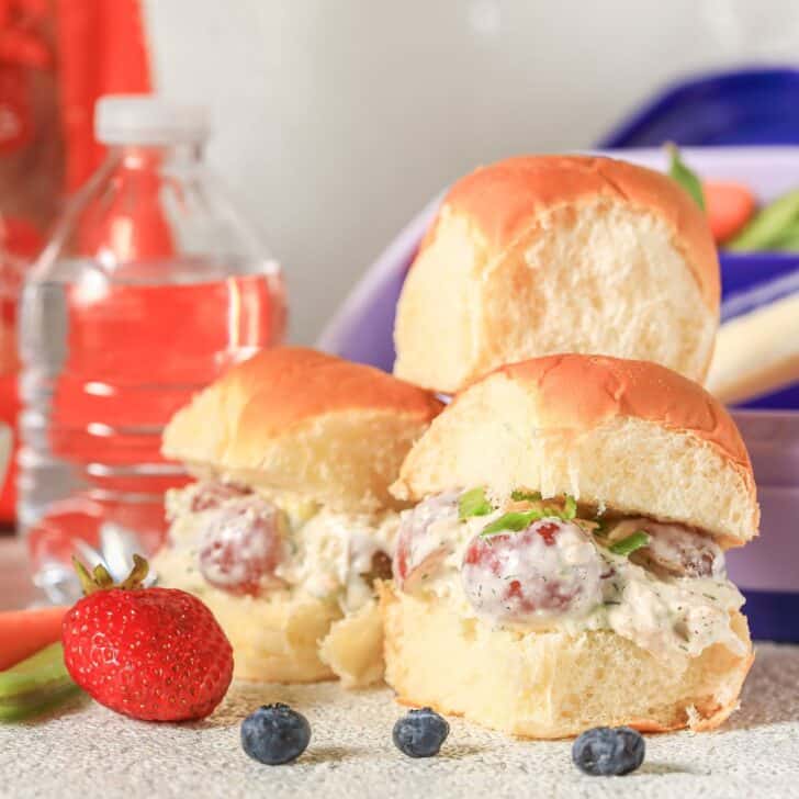 Celebrate With These Recipes for Lunch Ideas on Mother's Day Three chicken salad sliders with grapes and lettuce on bread rolls, arranged on a worktop with a strawberry, blueberries, a water bottle, and a purple container in the background.