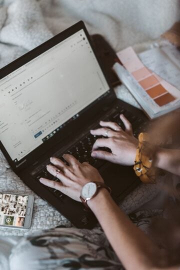 Person typing a safer family email on a laptop with a mobile displaying photos nearby, seated on a bed with a diary and colour swatches.