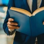 A person in a blue suit holding and reading an open Social Security Blue Book, with a blurred indoor background.