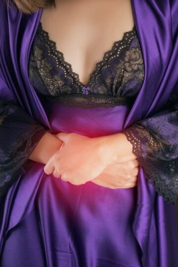 Woman in a purple dressing gown holding her abdomen with both hands, a red glow highlighting the stomach area, illustrating the importance of regular health checks for early detection in modern life.