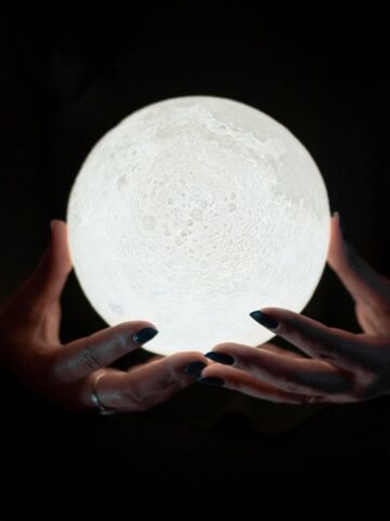 A person holds a glowing spherical lamp designed to look like the moon, evoking the mystery often associated with trusted psychic services, with both hands visible and a dark background.