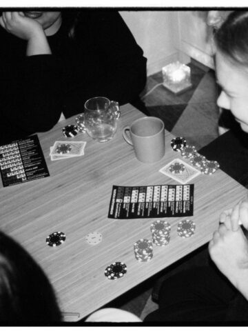 Four people are seated around a table playing a card game with poker chips, cards, drinks, and game instruction leaflets visible.