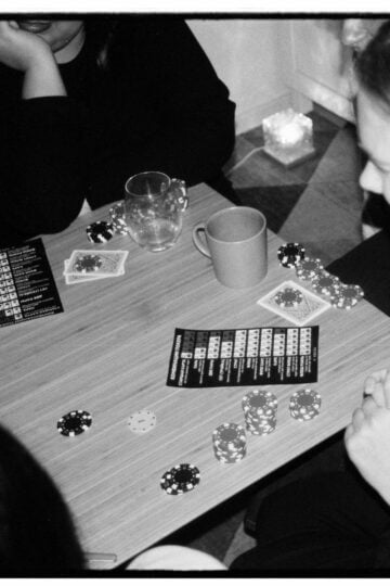Four people are seated around a table playing a card game with poker chips, cards, drinks, and game instruction leaflets visible.