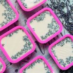 Square pink containers filled with a creamy white cold process soap, topped with dried lavender buds, are arranged on a patterned surface.