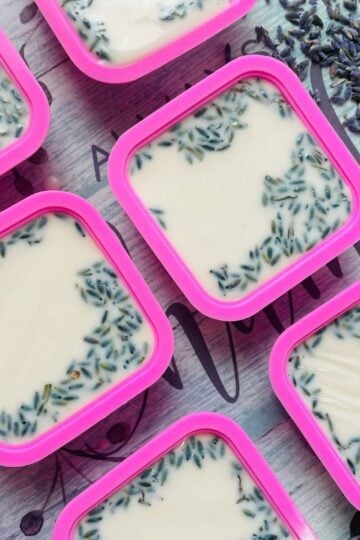 Square pink containers filled with a creamy white cold process soap, topped with dried lavender buds, are arranged on a patterned surface.