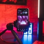 A smartphone on a tripod displays Instagram with a concert photo, capturing musicians in action, set in a dimly lit room glowing with red and blue lighting.