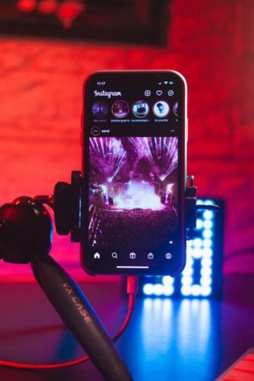 A smartphone on a tripod displays Instagram with a concert photo, capturing musicians in action, set in a dimly lit room glowing with red and blue lighting.
