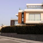 Modern two-storey house with high-performance windows, wooden accents, and glass balustrades, partially obscured by a tall hedge along a quiet street for comfort and style.