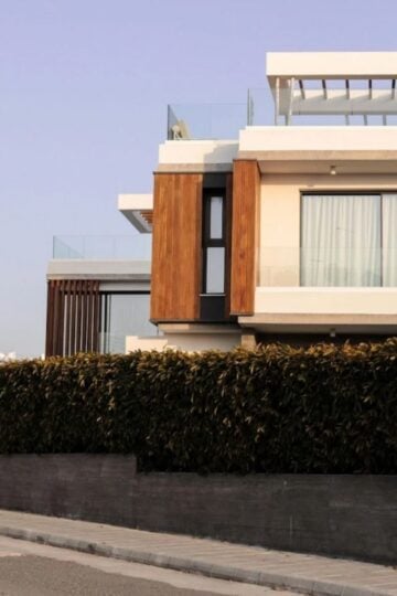 Modern two-storey house with high-performance windows, wooden accents, and glass balustrades, partially obscured by a tall hedge along a quiet street for comfort and style.
