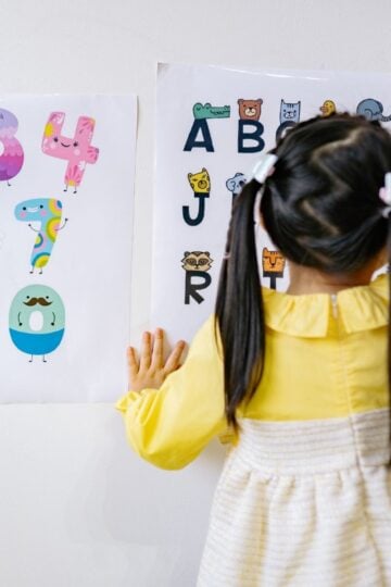 A young girl with plaits points at an alphabet chart on the wall, with a colourful number chart beside it—an ideal setup for nurseries using safe, hygienic wall sheets.