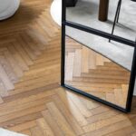 A rectangular mirror stands on parquet flooring in a herringbone pattern, partially reflecting a beige rug and the leg of a black chair, highlighting the elegant flooring style.