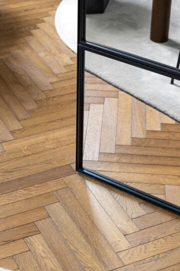A rectangular mirror stands on parquet flooring in a herringbone pattern, partially reflecting a beige rug and the leg of a black chair, highlighting the elegant flooring style.
