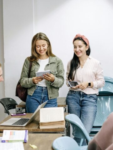 Four students stand and sit around a desk with notebooks, a laptop, and papers in a classroom, engaging in conversation during a group study session.