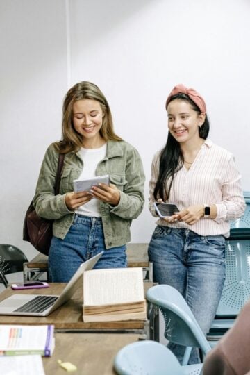 Four students stand and sit around a desk with notebooks, a laptop, and papers in a classroom, engaging in conversation during a group study session.