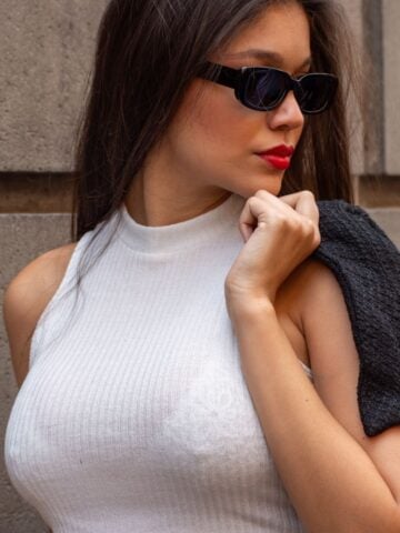 A woman wearing a sleeveless white polo-neck top, black sunglasses, and red lipstick holds a dark jacket over her shoulder while standing against a stone wall, exuding confidence reminiscent of a breast uplift transformation.