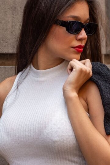 A woman wearing a sleeveless white polo-neck top, black sunglasses, and red lipstick holds a dark jacket over her shoulder while standing against a stone wall, exuding confidence reminiscent of a breast uplift transformation.