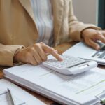A person in business attire uses a calculator and laptop whilst reviewing financial documents on a desk, preparing for the 2026 launch of a Stripe-like payment system.