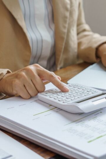 A person in business attire uses a calculator and laptop whilst reviewing financial documents on a desk, preparing for the 2026 launch of a Stripe-like payment system.
