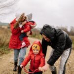 A family makes the most of a cloudy weekend outdoors; a woman holds a child whilst a man helps another child ride a small red bicycle.