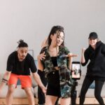 Three people dance in front of a mobile on a tripod indoors, possibly filming a TikTok video; one woman is centred, flanked by two others in casual clothing-perhaps hoping to earn more TikTok Likes.