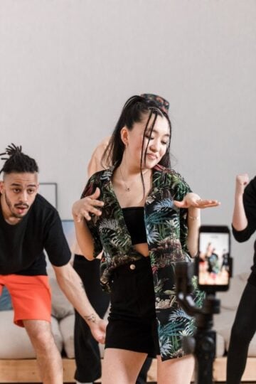 Three people dance in front of a mobile on a tripod indoors, possibly filming a TikTok video; one woman is centred, flanked by two others in casual clothing—perhaps hoping to earn more TikTok Likes.