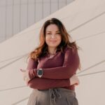 Woman with long brown hair wearing a maroon top and smartwatch stands with arms crossed, smiling in front of a modern light-coloured building-demonstrating how small changes can boost your confidence every day.