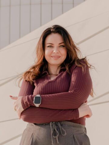 Woman with long brown hair wearing a maroon top and smartwatch stands with arms crossed, smiling in front of a modern light-coloured building-demonstrating how small changes can boost your confidence every day.