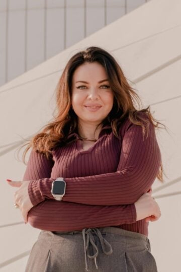 Woman with long brown hair wearing a maroon top and smartwatch stands with arms crossed, smiling in front of a modern light-coloured building—demonstrating how small changes can boost your confidence every day.