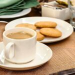 A white cup of coffee sits on a saucer next to a plate with four biscuits, perfect for dunking-a beloved British tradition-on a brown placemat. Serviettes and sugar sachets are in the background.