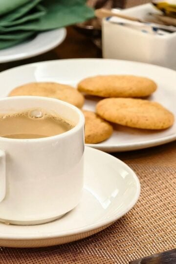 A white cup of coffee sits on a saucer next to a plate with four biscuits, perfect for dunking—a beloved British tradition—on a brown placemat. Serviettes and sugar sachets are in the background.