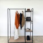 A minimalist open wardrobe-perfect for busy mums-features four hanging garments, a hat, black handbag, jewellery, and black shoes on shelves against a white wall for an effortless capsule wardrobe approach.
