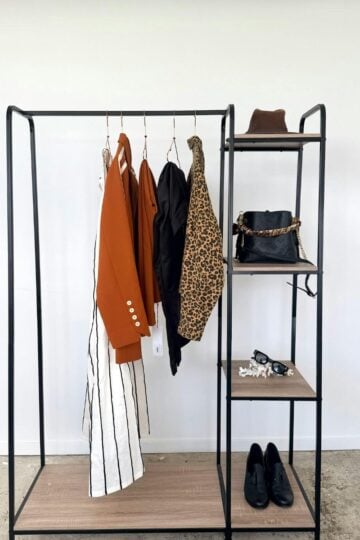 A minimalist open wardrobe—perfect for busy mums—features four hanging garments, a hat, black handbag, jewellery, and black shoes on shelves against a white wall for an effortless capsule wardrobe approach.