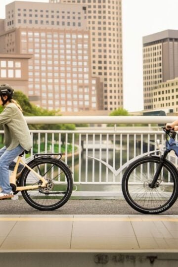 Two people wearing helmets ride electric bicycles on a bridge with city buildings and trees in the background, showcasing versatile riding scenarios for any electric bike selection.