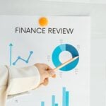 A person points a pen at a pie chart labelled "FINANCE REVIEW" on a paper with financial data, highlighting currency percentages and strategies for website publishers seeking sustainable revenue streams.
