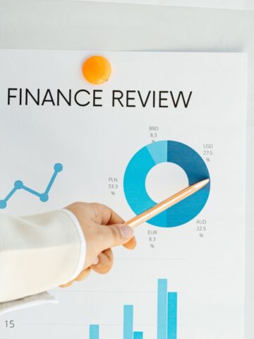 A person points a pen at a pie chart labelled "FINANCE REVIEW" on a paper with financial data, highlighting currency percentages and strategies for website publishers seeking sustainable revenue streams.