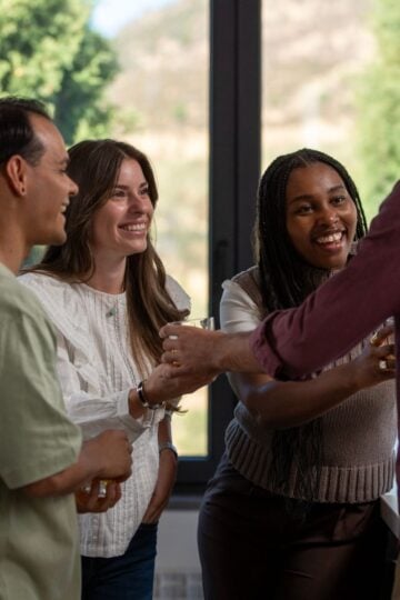 Four people stand indoors near large windows, holding drinks and smiling as they engage in conversation. Trees and a hilly landscape are visible outside.