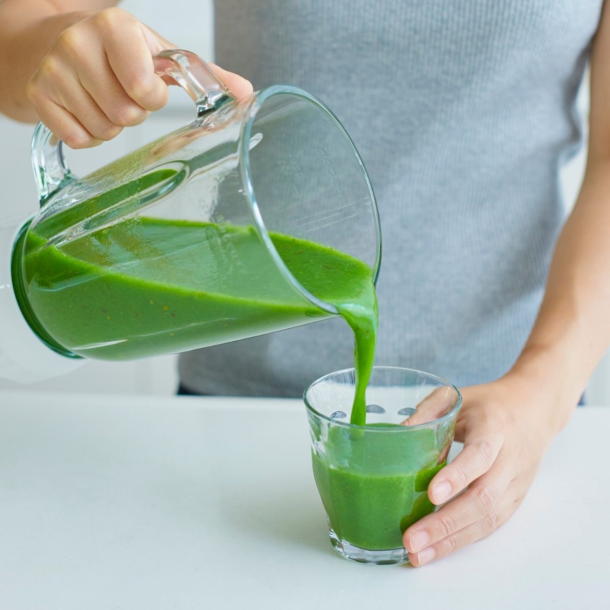 A person pours a thick green smoothie from a blender jug into a glass on a white surface, showing one way how to take collagen peptides and enjoy their benefits in your daily routine.