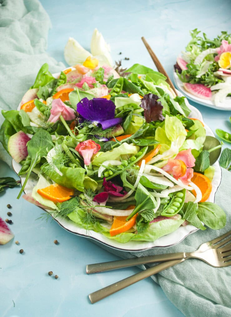 A plate of mixed green salad with orange slices, mangetout, radish, fennel, edible flowers, and fresh herbs, set on a blue surface with a fork and knife beside it.
