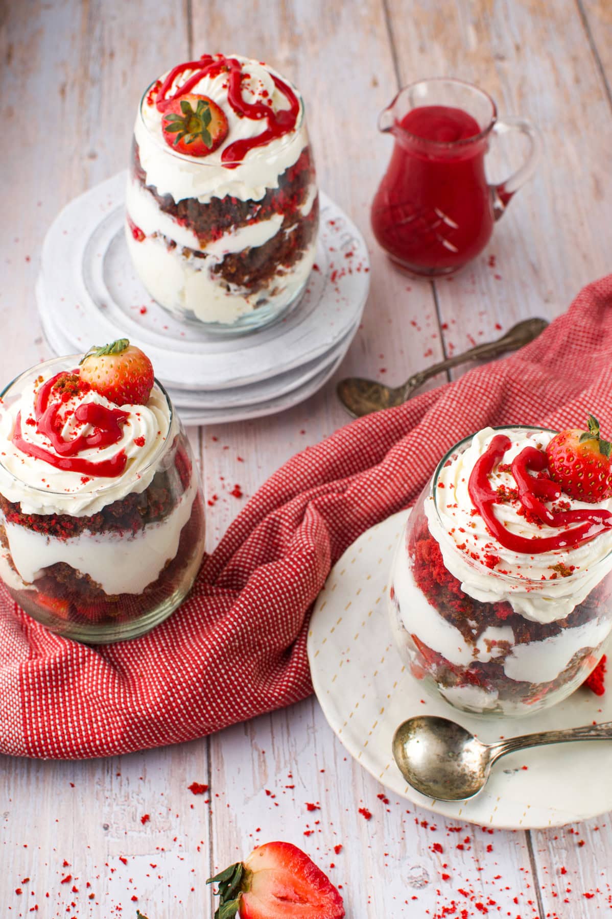 Three glass jars filled with layered strawberry and cream dessert sit on plates, garnished with strawberries and red sauce, with a jug of sauce and a spoon nearby on a wooden table.