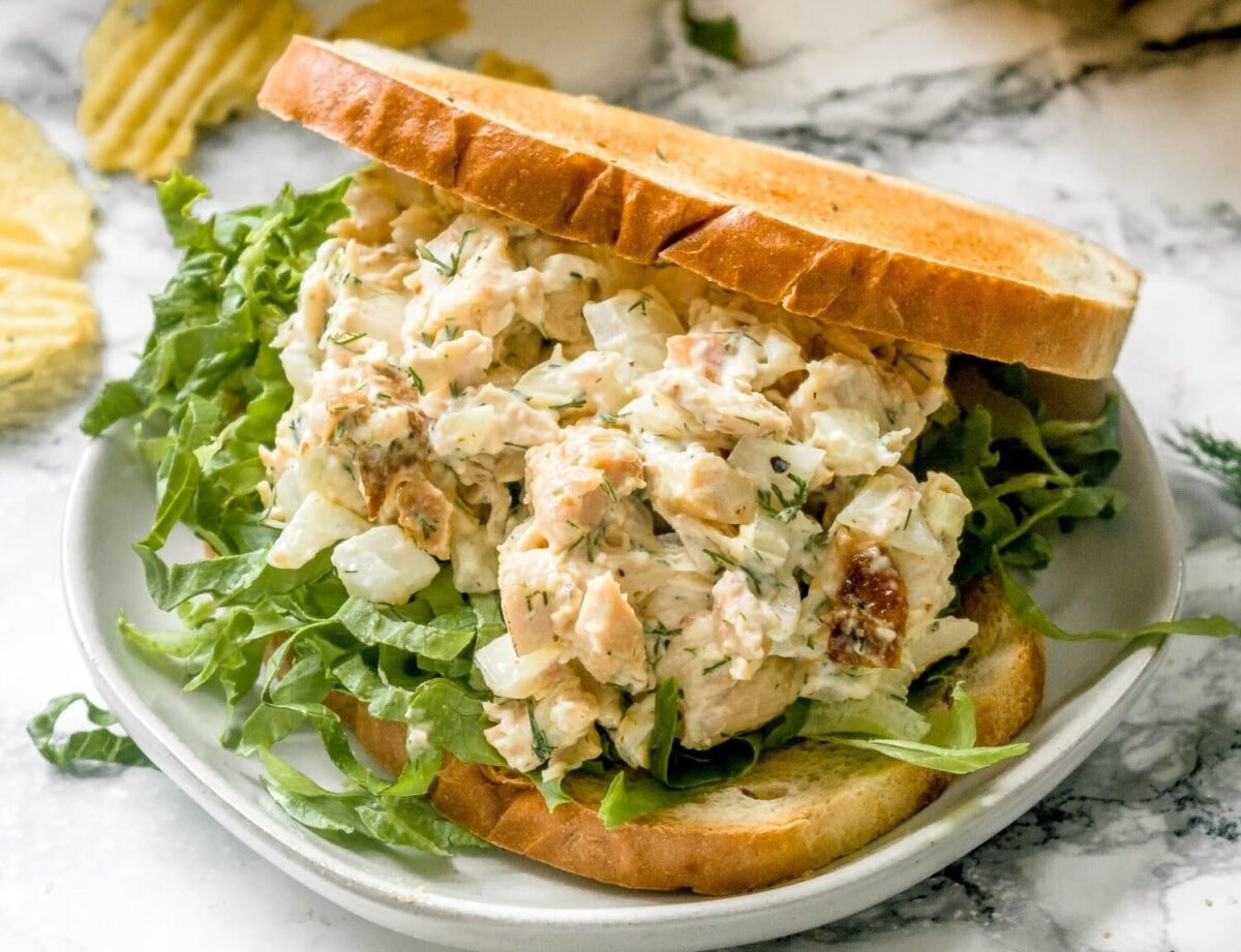 Chicken salad with chopped onions and herbs on green lettuce between two slices of toasted bread, served on a plate with crisps in the background.