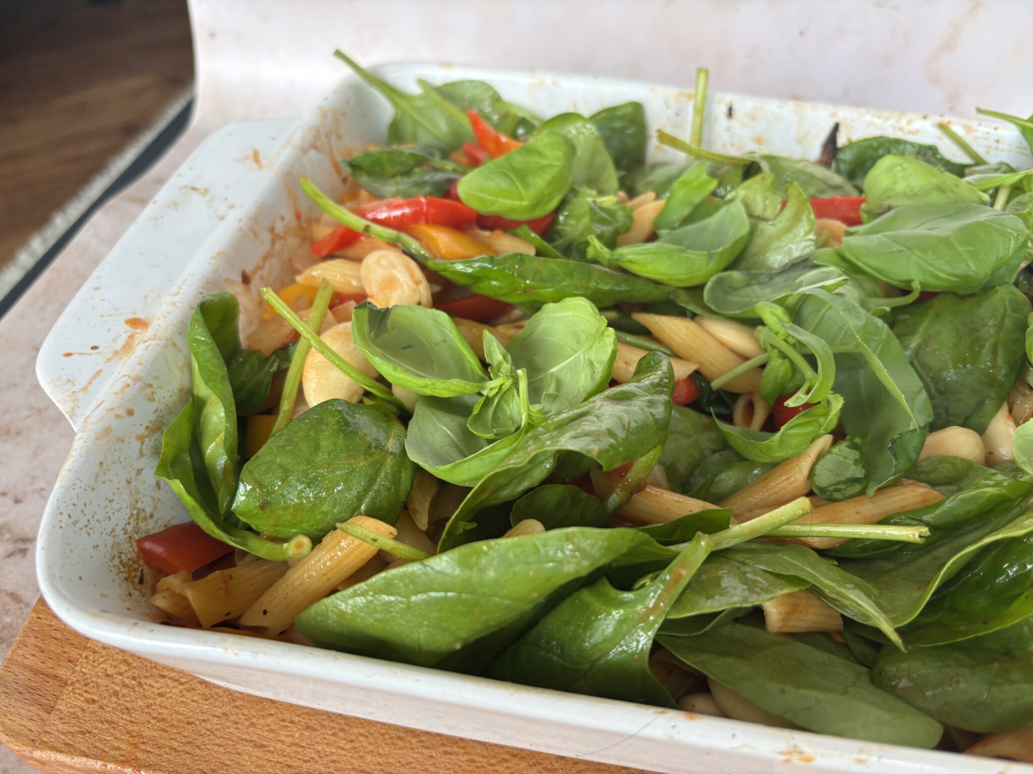 A white baking dish filled with penne pasta, red peppers, cashew nuts, and topped with fresh spinach leaves-perfect for Mother's Day or as inspiration for your next lunch ideas.