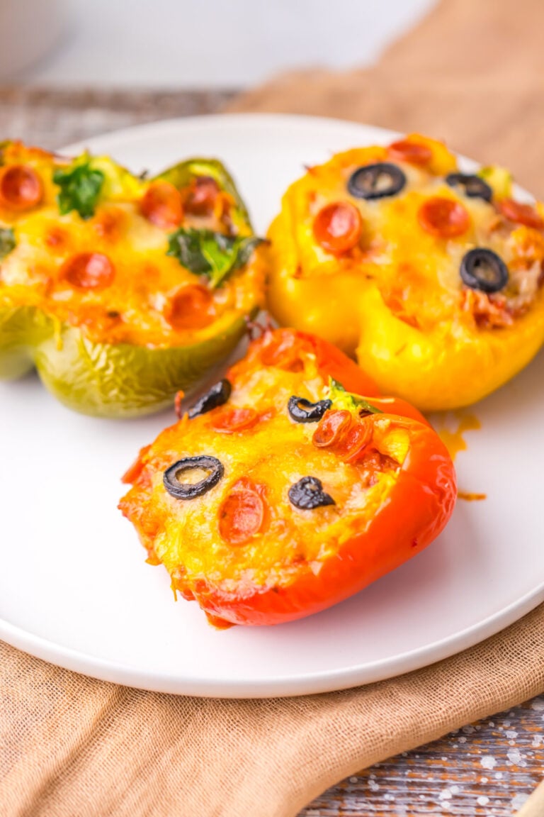 Three stuffed pepper halves topped with melted cheese, sliced black olives, mini pepperoni, and herbs are arranged on a white plate.