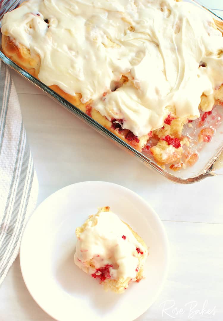 A piece of iced cherry pudding is served on a white plate next to a glass baking dish with the rest of the pudding-like dessert.