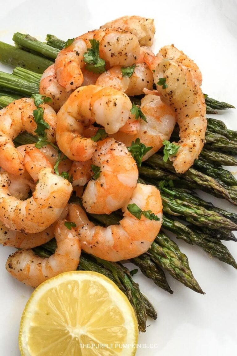 Cooked prawns garnished with herbs served on roasted asparagus with a wedge of lemon on a white plate.