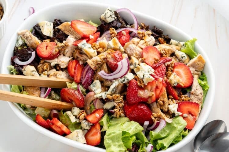 A salad bowl with lettuce, grilled chicken, strawberries, red onions, blue cheese, and granola, with wooden salad servers.