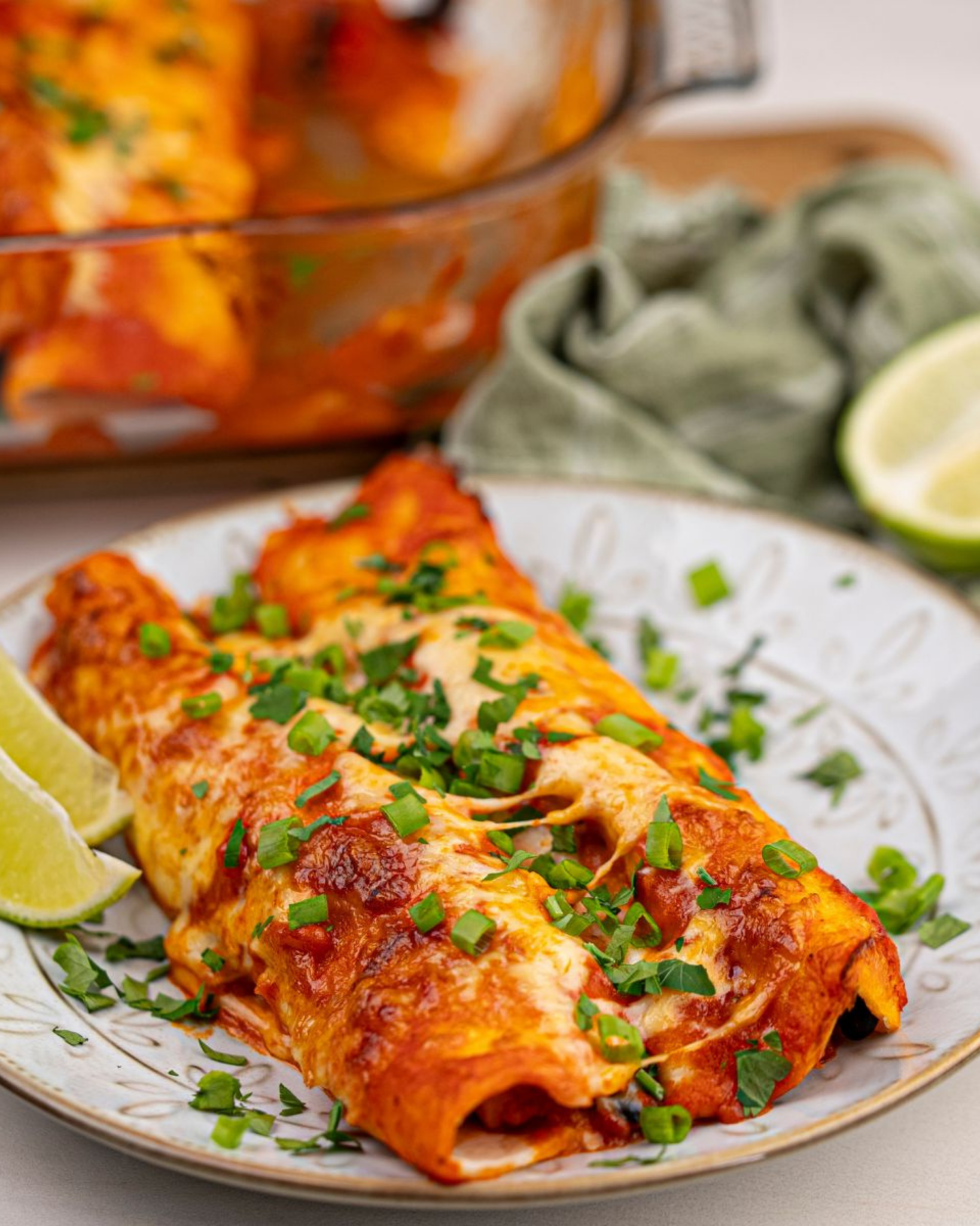 Celebrate With These Recipes for Lunch Ideas on Mother's Day Two cheesy enchiladas topped with chopped spring onions are served on a plate with lime wedges-perfect for lunch ideas or Mother's Day lunch recipes-with a baking dish and a green cloth in the background.