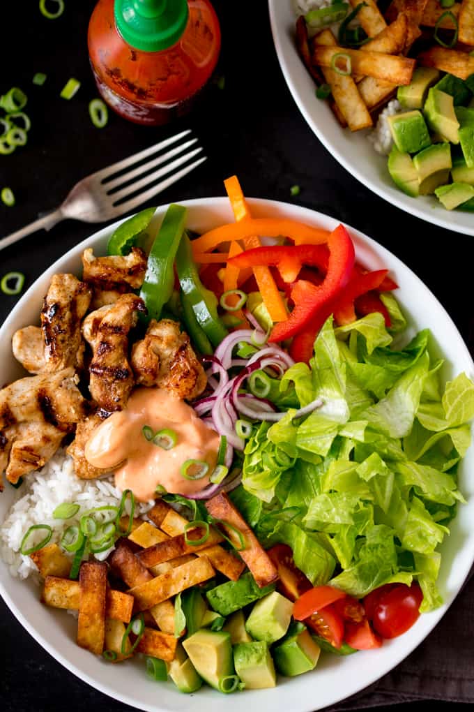 Celebrate With These Recipes for Lunch Ideas on Mother's Day A bowl with grilled chicken, rice, lettuce, avocado, peppers, onions, tomatoes, fried tofu, sliced spring onions, and sauce, with a bottle of sriracha and a fork nearby.