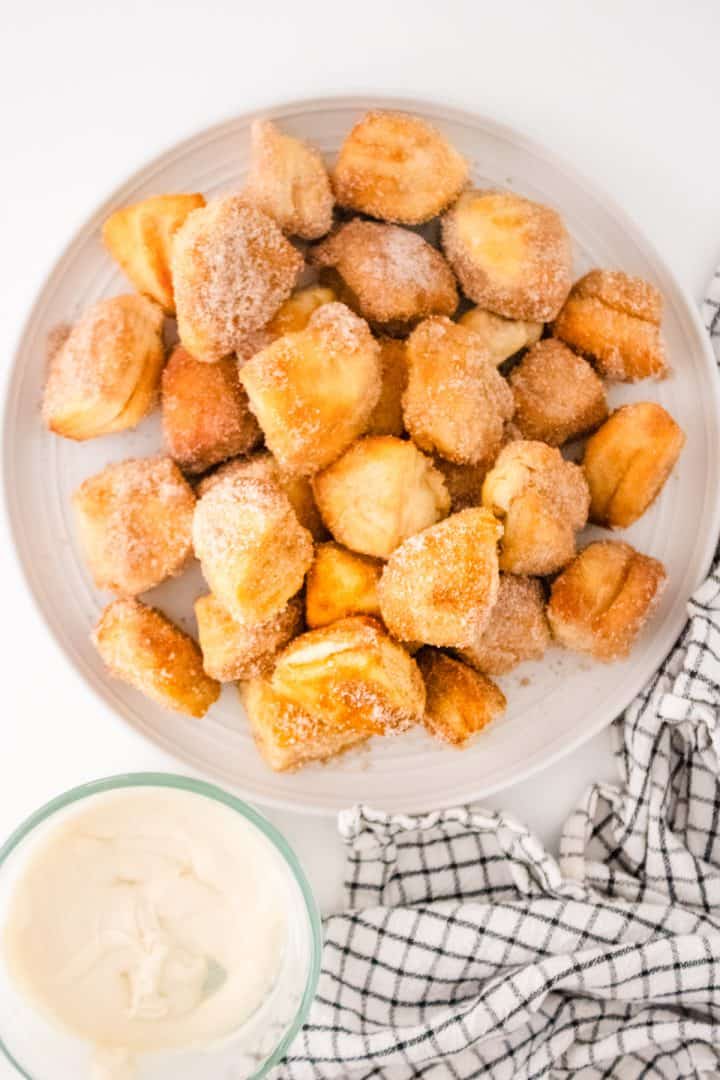 Plate of cinnamon sugar-coated biscuit bites with a small bowl of white dipping sauce and a black-and-white chequered cloth nearby.