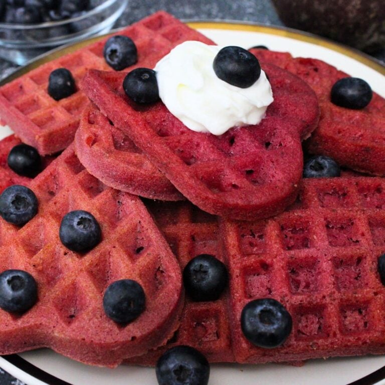 A plate of heart-shaped red waffles topped with blueberries and a dollop of cream.